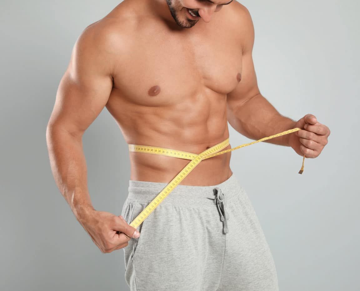 Young man with slim body using measuring tape on grey background